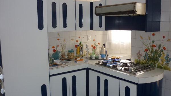 Cozinha com armários embutidos, forno embutido, pequena mesa para lanches e área externa com mais uma geladeira e dispensa.